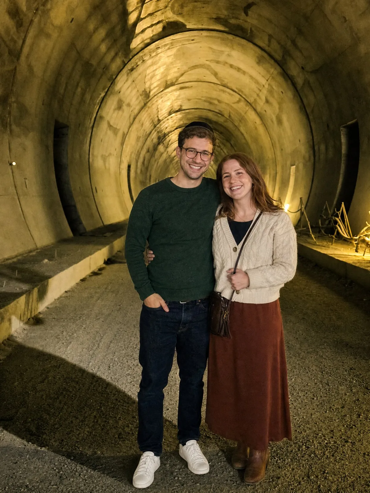 Ein Paar in einem beleuchteten Betontunnel: er trägt eine schwarze Kippa, Brille, grünen Pullover und Jeans; sie trägt eine cremefarbene Strickjacke und einen rostroten Rock