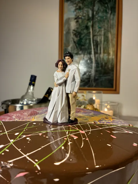 Printed figure of a bride in a white gown and a groom in a white jacket on top of a chocolate cake with colored drizzle