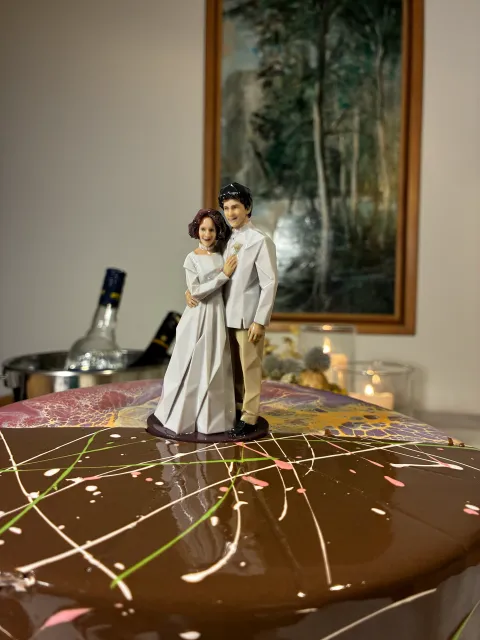 Glossy faceted figure of a bride in a long white gown and a groom in a white jacket and cream trousers, holding each other, on a chocolate glazed cake