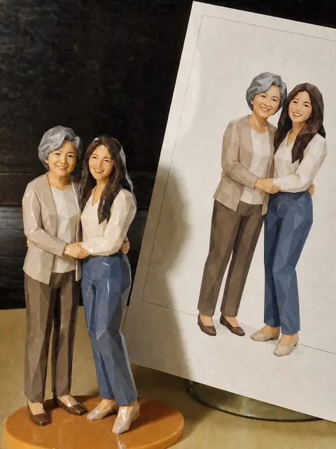 Glossy faceted figure of a gray-haired mother in a beige jacket and her grown daughter in a white blouse and jeans, hugging on a round orange base, beside a card showing the same pair.
