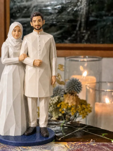 Topper ng bride na naka-puting hijab at long-sleeved na white gown na nakahawak sa braso ng groom na naka-ivory na sherwani, sa navy na base sa glazed cake, may kandila sa likod.