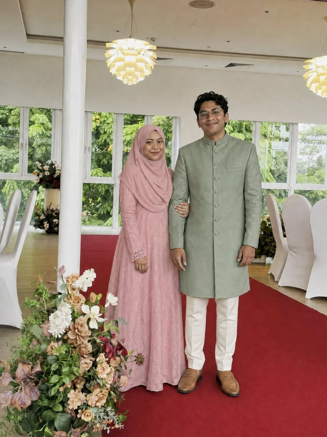 A bride in a dusty-rose hijab and long dress beside a groom in a sage-green sherwani and cream trousers, on a red carpet aisle in a bright hall.