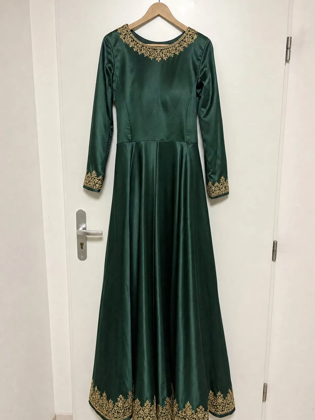 A long-sleeved emerald green dress with gold embroidery at the neck, cuffs and hem, on a wooden hanger on a white door.