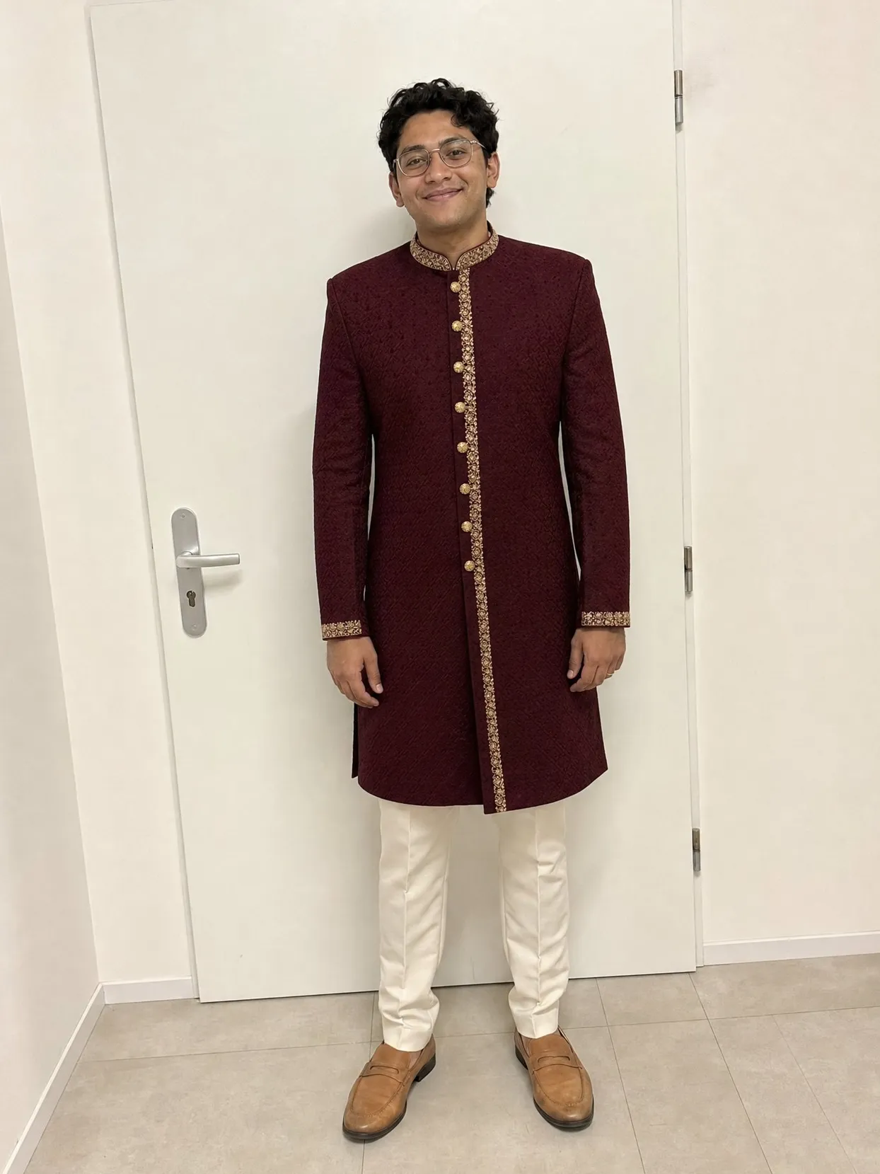 The groom from the first photo in a maroon sherwani with gold trim and ivory trousers, in front of a white door.
