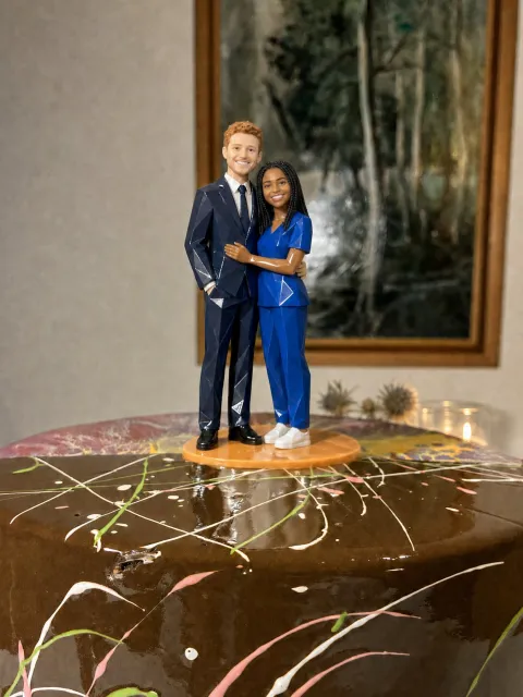 Couple topper sa orange na base sa ibabaw ng chocolate cake. Mahaba ang tirintas ng bride at naka-royal blue na scrubs at puting sneakers. Naka-navy na suit ang groom na pula ang buhok.