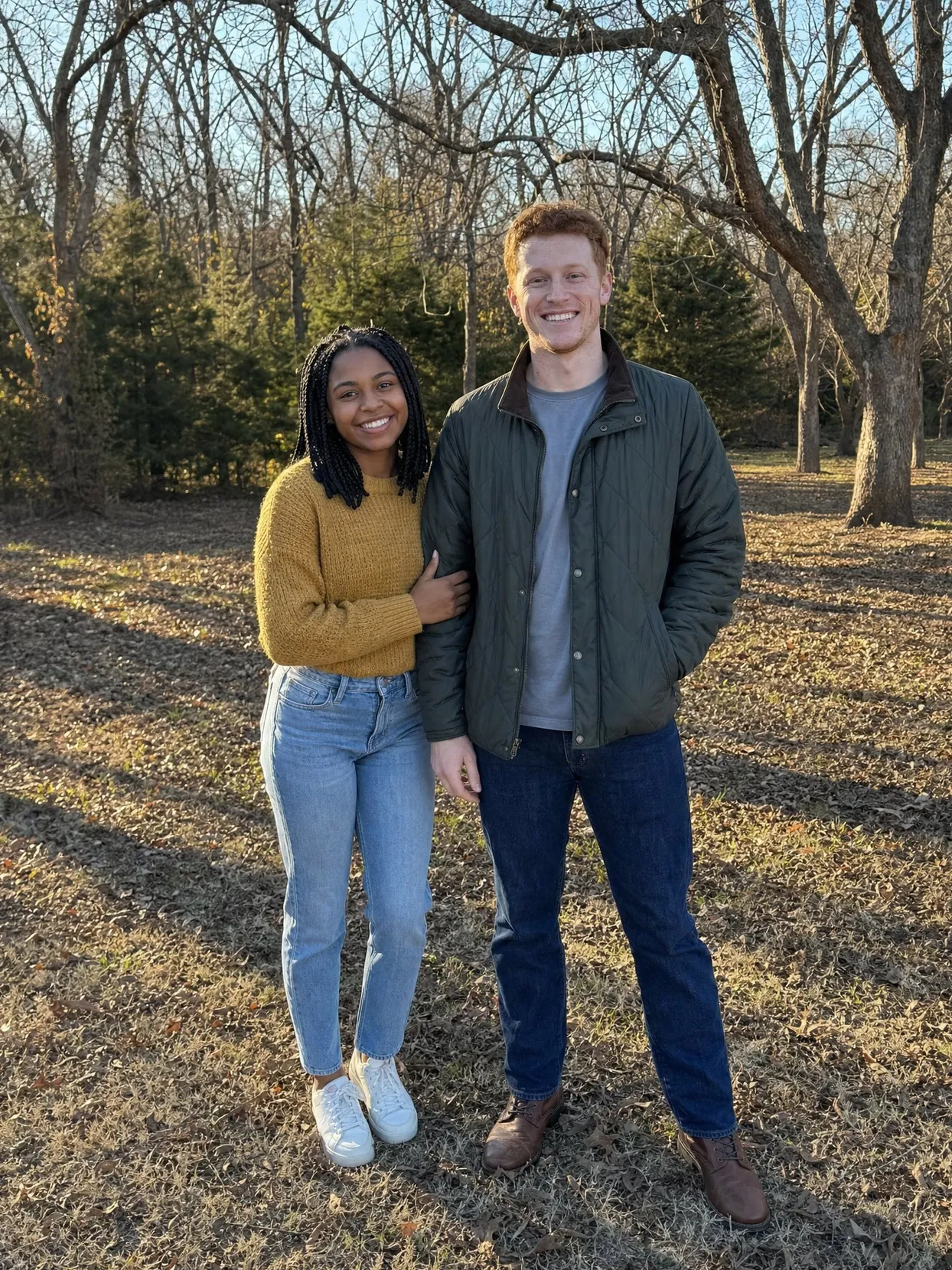 A woman in a mustard sweater and jeans and a man in a green jacket standing on winter grass under bare trees.
