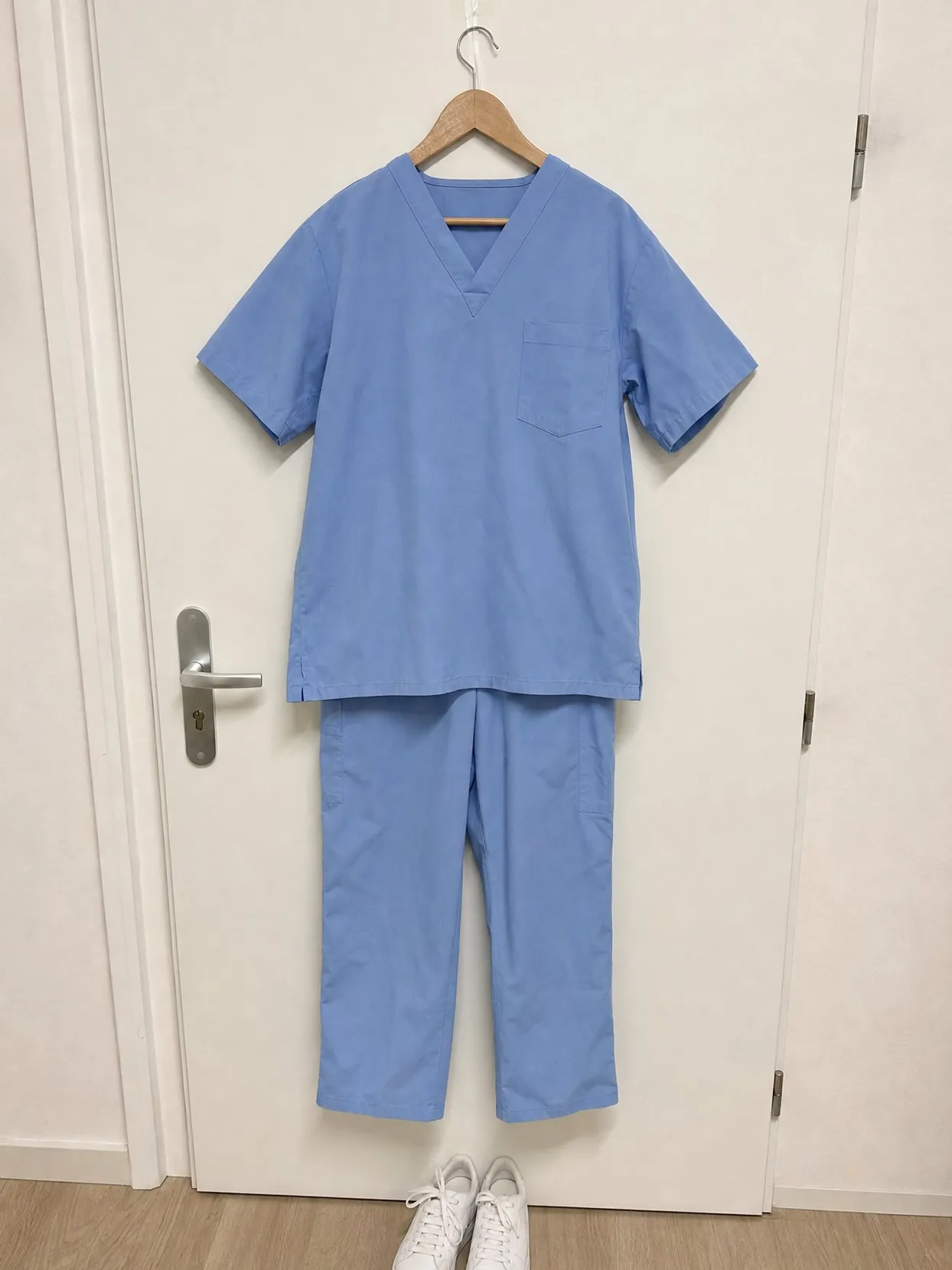 Light blue scrub top and pants on a hanger on a white door, white sneakers below.