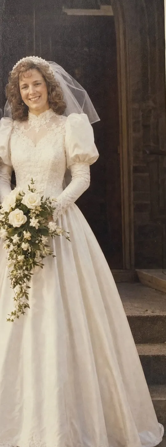 Old color snapshot of a young bride with permed curls and a short veil, in a white satin gown with puffed sleeves, holding white roses.