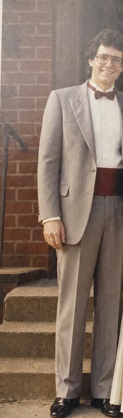 Old color snapshot of a young groom with dark curly hair and large glasses, in a pale gray tuxedo with a burgundy bow tie.
