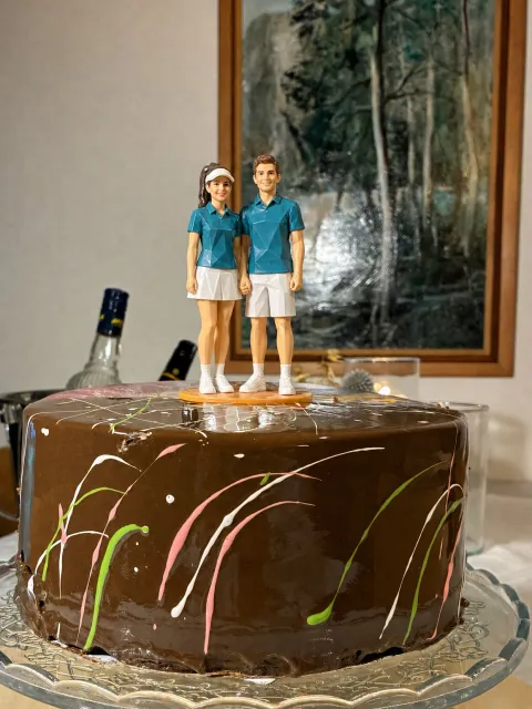 Origami topper ng couple na naka-magkaparehong teal na polo, siya naka-puting skort at visor, siya naman naka-puting shorts, magkahawak-kamay, sa ibabaw ng chocolate drip cake.