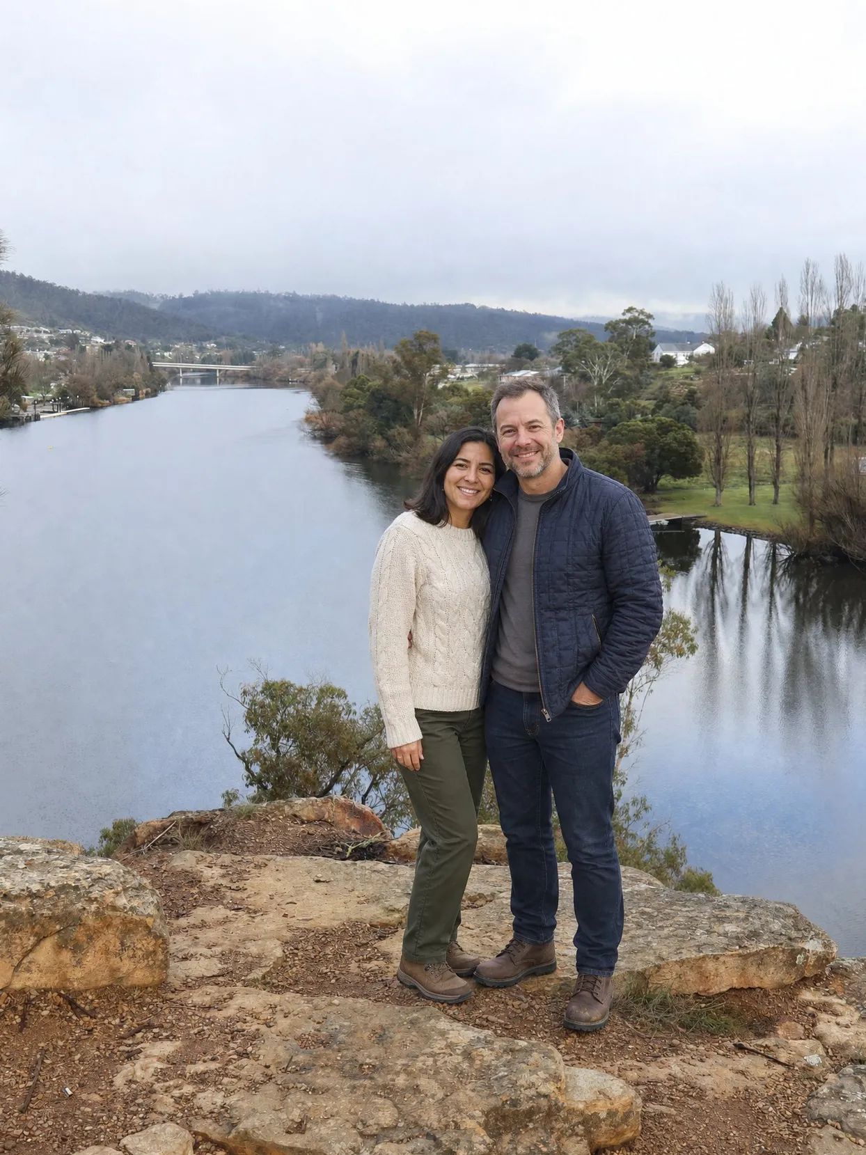 A couple at a river lookout, she in a cream sweater and green trousers, he in a navy quilted jacket and jeans.