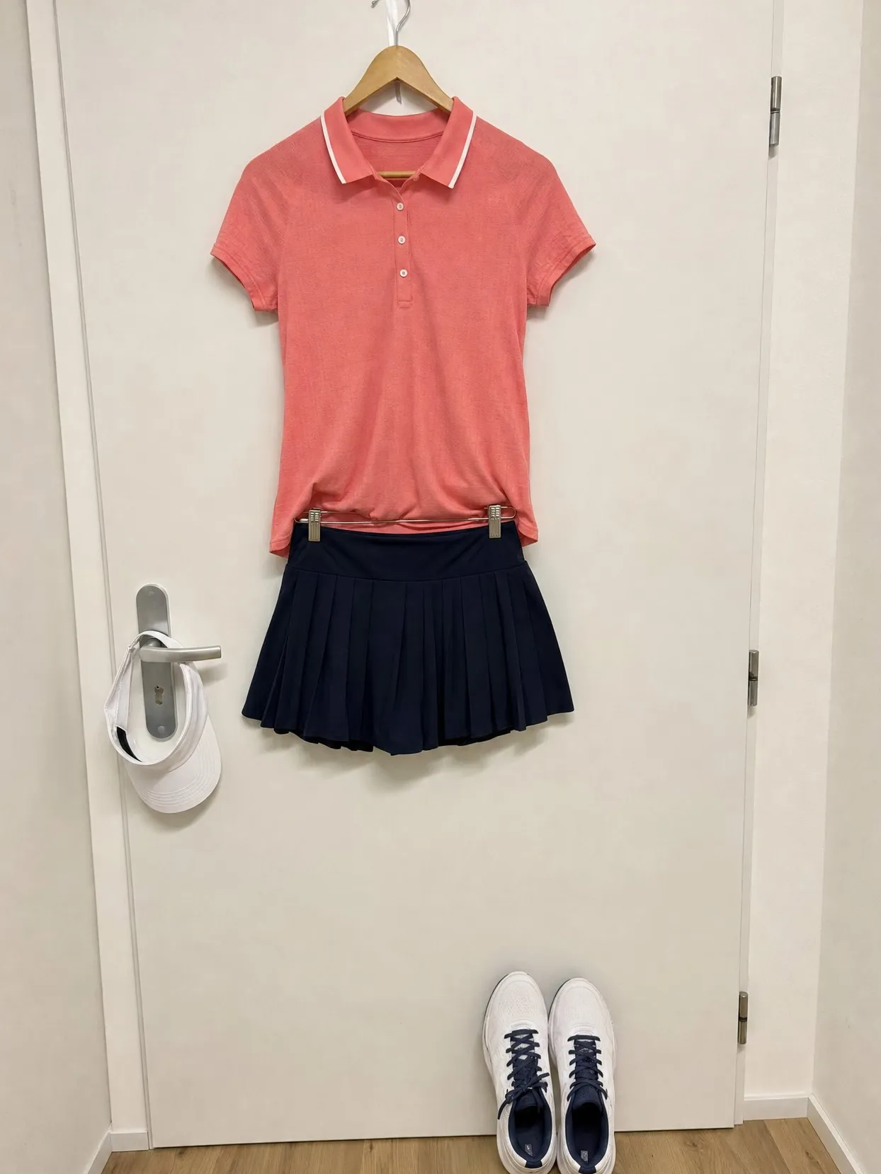 A coral polo with a white collar edge and a navy pleated skort on a hanger, a visor on the handle and white shoes below.