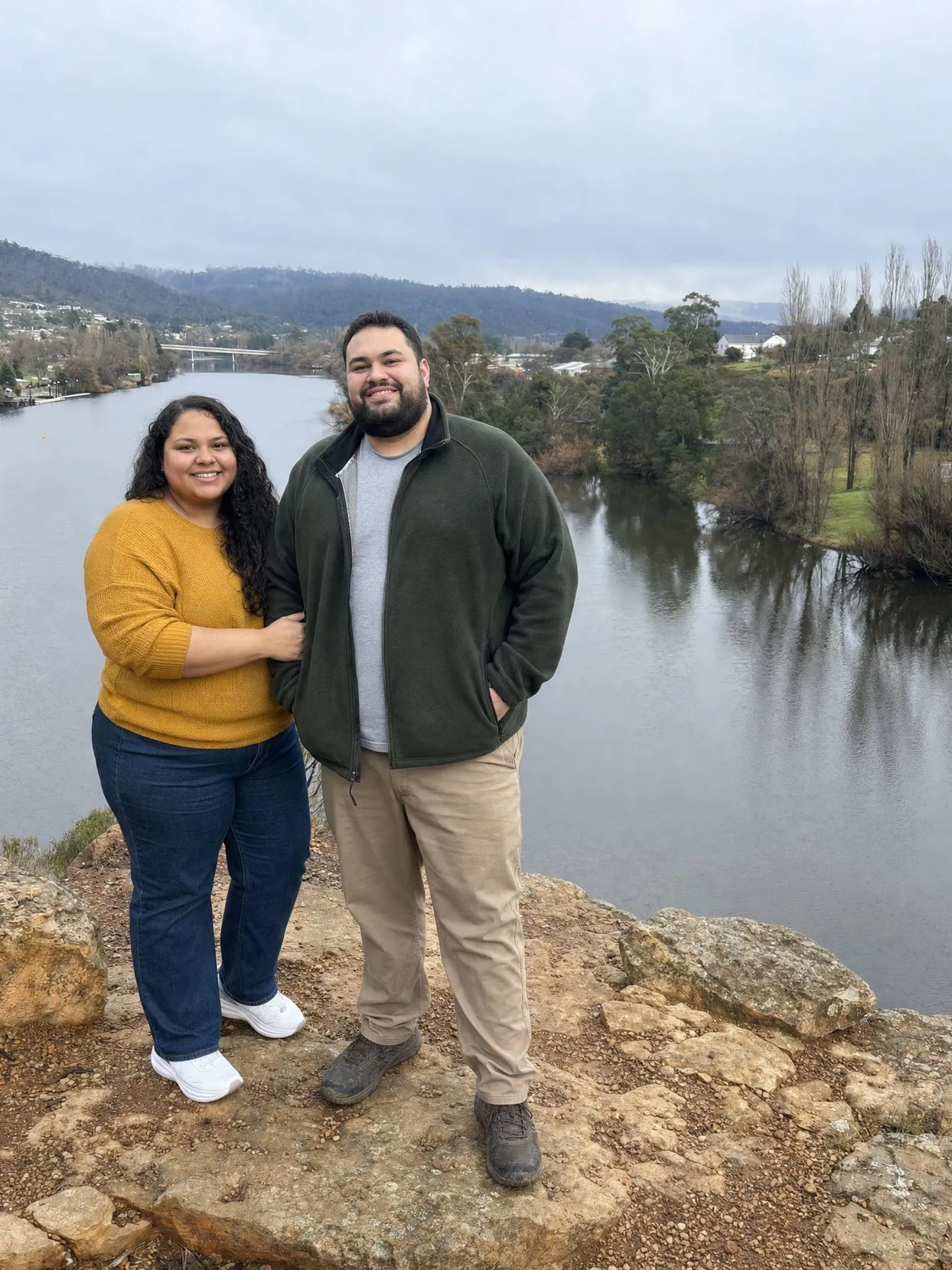 A plus size woman in a mustard sweater and jeans and a heavy-set man in a green fleece and khakis stand side by side on a rock above a river.