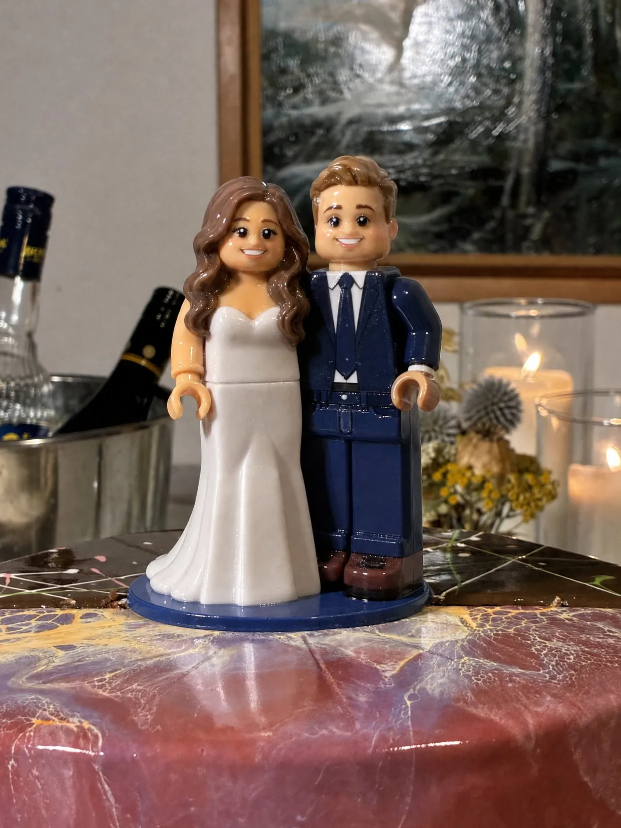 Brick Figures cake topper of a bride with long brown hair in a white gown and a groom in a navy suit, on a blue base on a marbled pink cake.