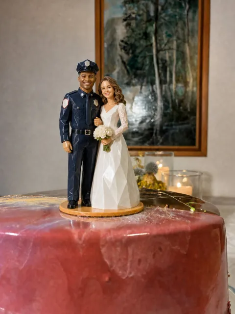 Couple topper sa pink na glazed cake. Naka-navy na police uniform siya na may kurbata, plain na badge at cap. Naka-puting gown naman siya na may lace na manggas at may hawak na bouquet.