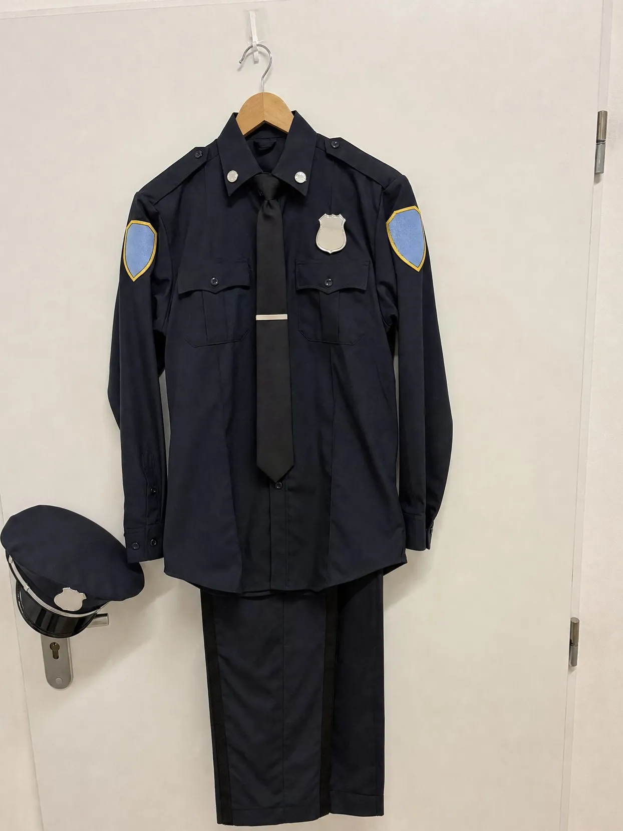 A navy uniform shirt with a black tie, a tie bar and light-blue patches with gold borders on a hanger on a white door, the cap on the door handle.