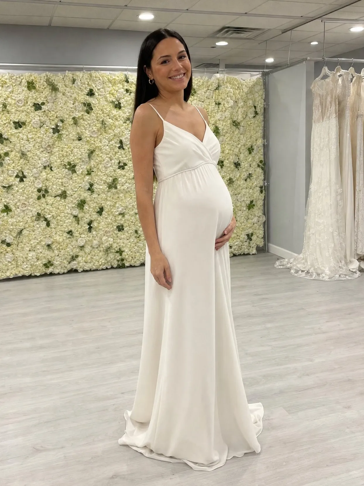 Dieselbe Frau in einem elfenbeinfarbenen Empire-Kleid in einem Brautmodengeschäft, seitlich gedreht, sodass ihr Babybauch von der Seite zu sehen ist