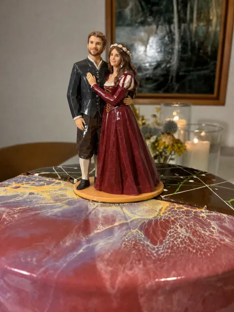 Cake topper sa marbled na pink at chocolate cake: bride na naka-deep red na gown na may laced bodice, puffed sleeves at flower wreath, at groom na naka-charcoal na doublet, brown na breeches at cream na stockings.