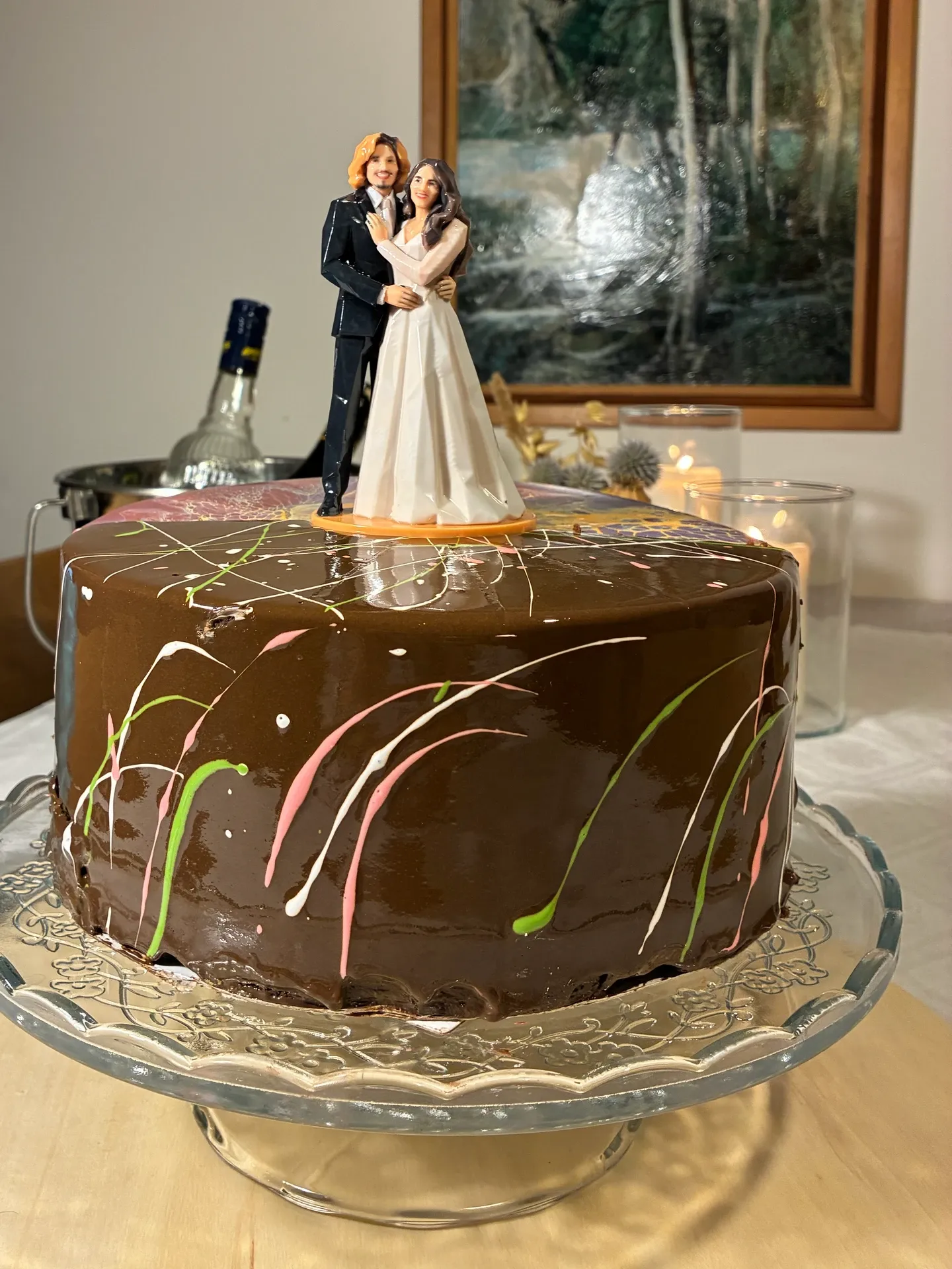 Bride and groom figure centered on a one-tier chocolate cake on a glass stand
