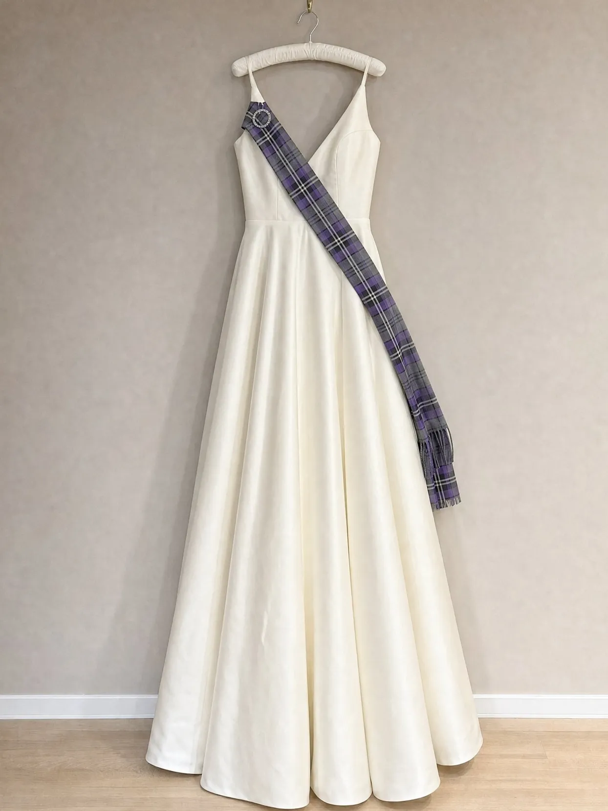 An ivory wedding gown on a padded hanger on a wall hook, a gray and purple tartan sash pinned at one shoulder with a silver brooch