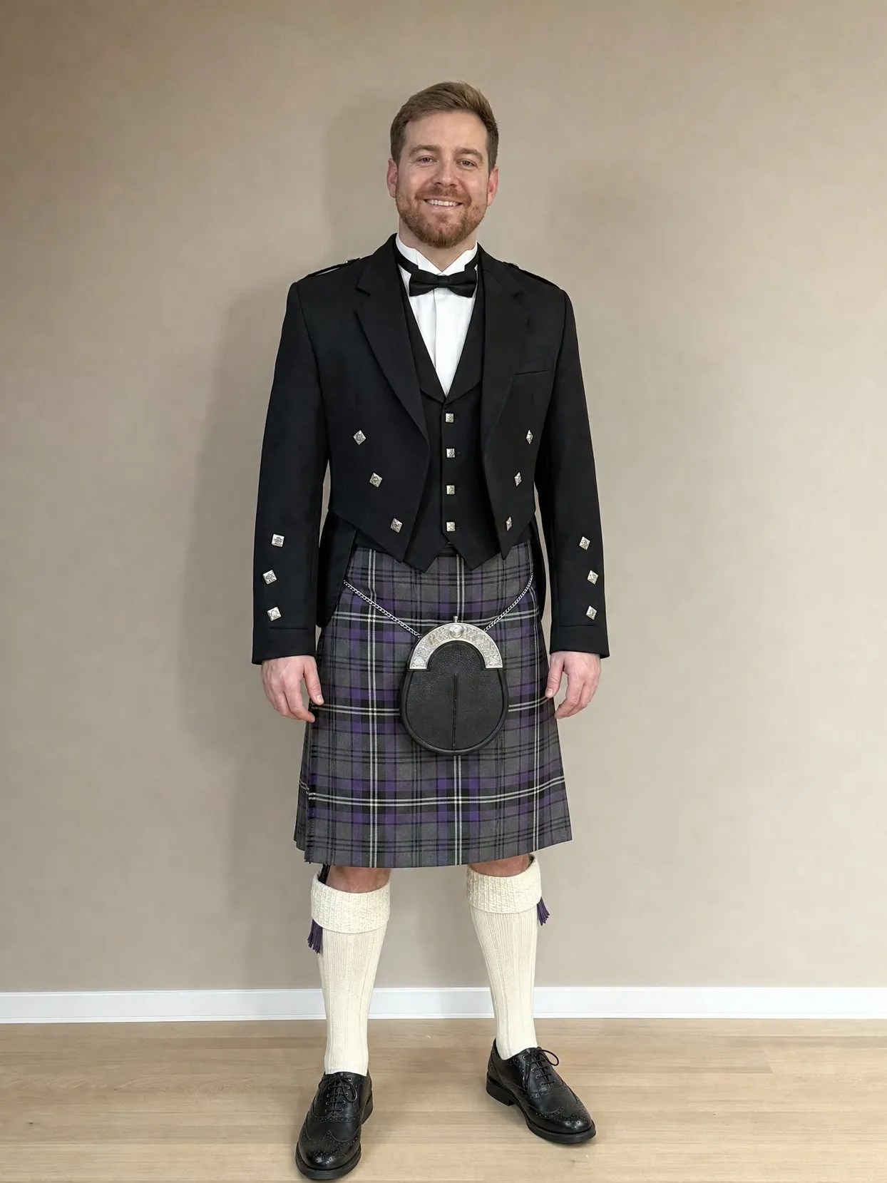 The same man standing against a plain wall in a gray and purple tartan kilt, black Prince Charlie jacket, bow tie, black sporran, cream kilt socks and black brogues
