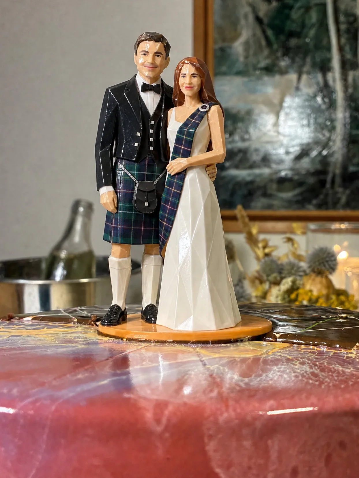 Origami cake topper of a groom in a navy and green tartan kilt, black Prince Charlie jacket, waistcoat and bow tie, beside a bride in white with a matching tartan sash, on a pink glazed cake