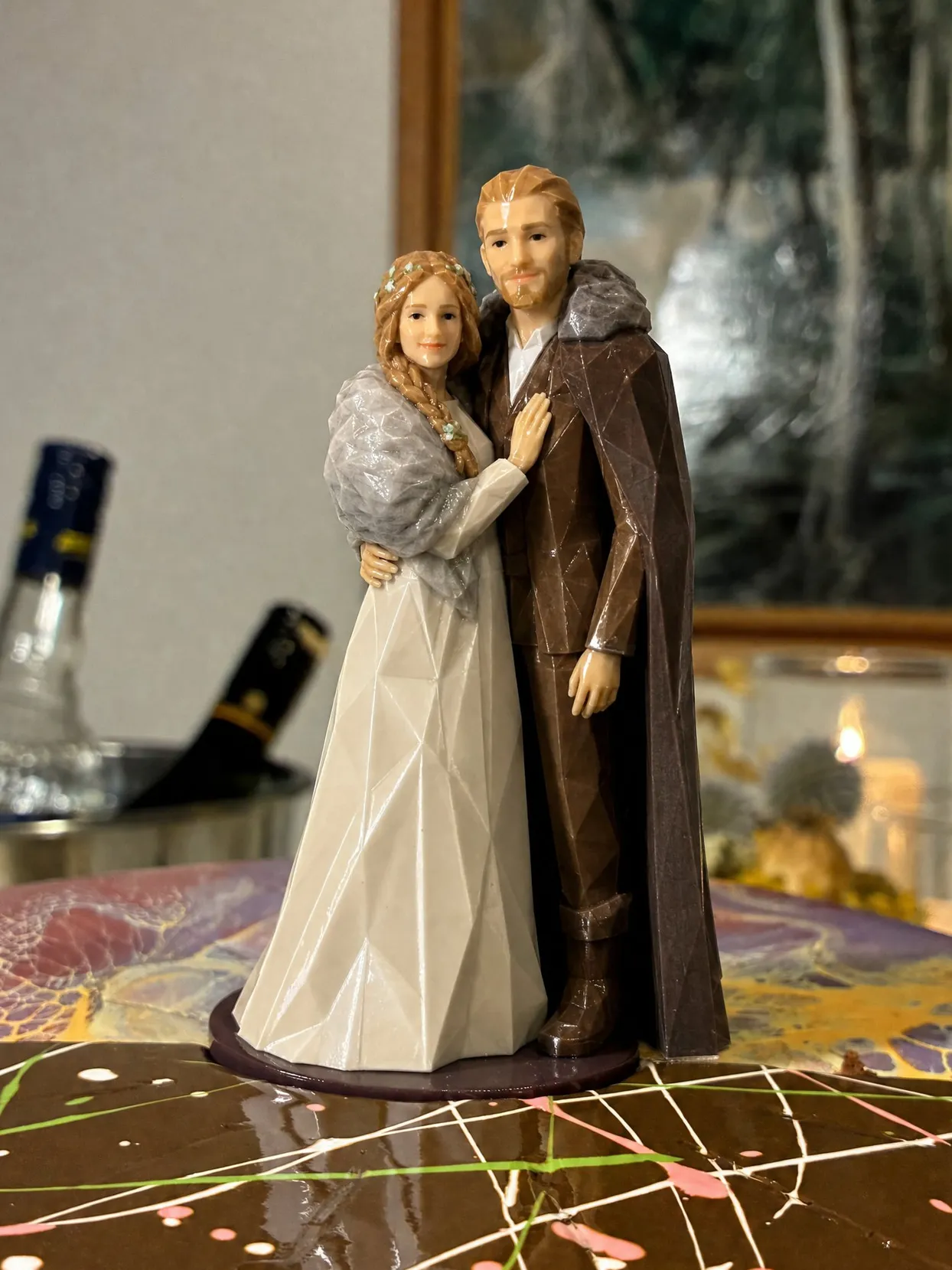 A couple topper on a chocolate cake, the bride in an ivory gown with a grey fur wrap and a braided flower crown, the groom in a brown suit and a cloak with a fur collar