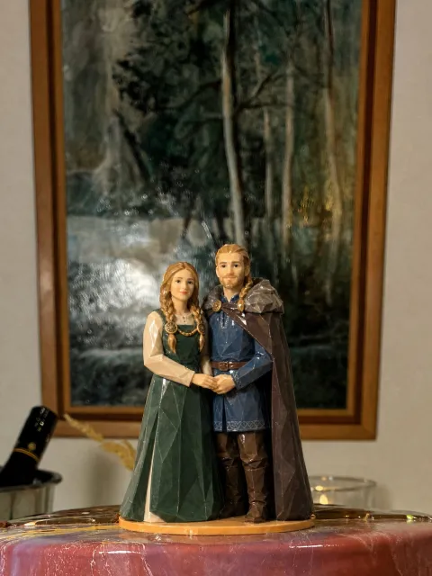 Isang couple topper na naka-Viking na damit pangkasal at magkahawak-kamay sa pulang wedding cake, siya na naka-green apron dress na may bronze brooch at tirintas, siya na naka-blue tunic at brown cloak na may fur collar
