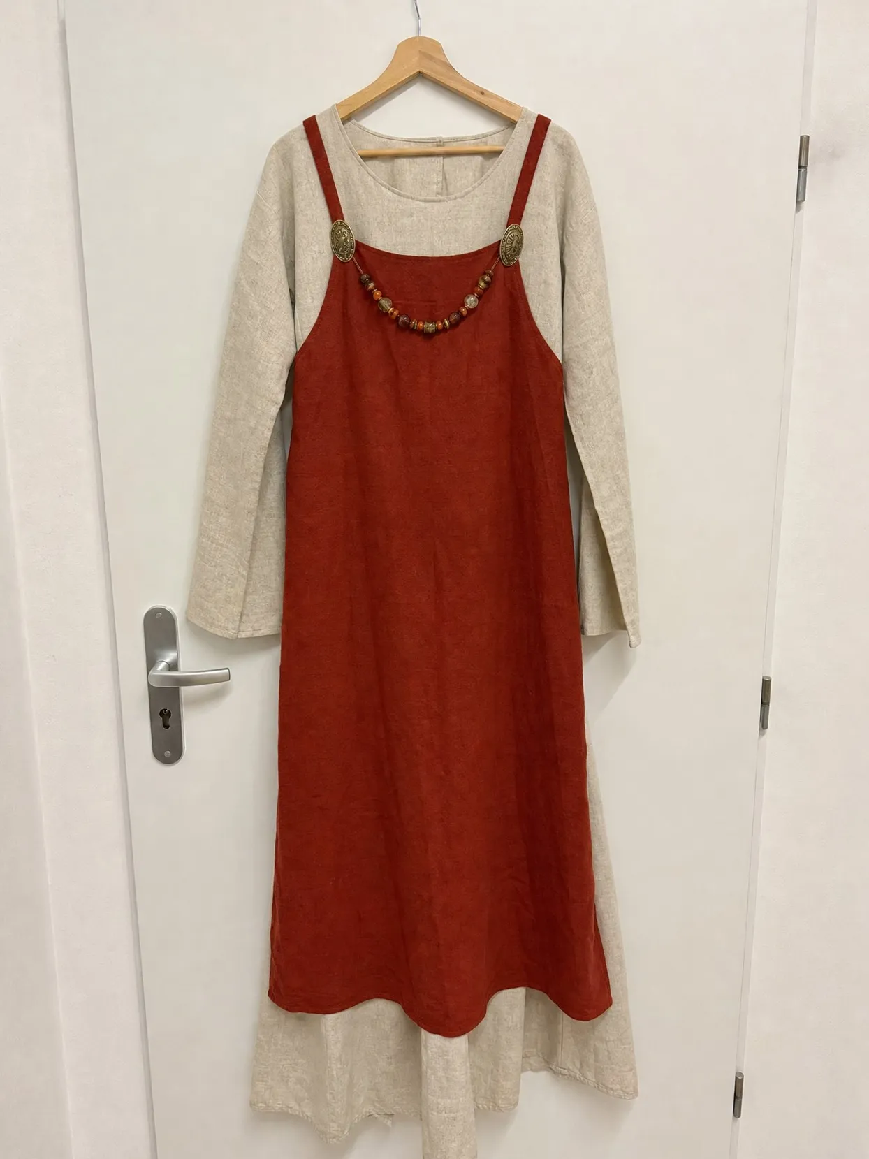 A rust-red apron dress with two oval bronze brooches and a string of beads over a long linen underdress, on a wooden hanger on a white door