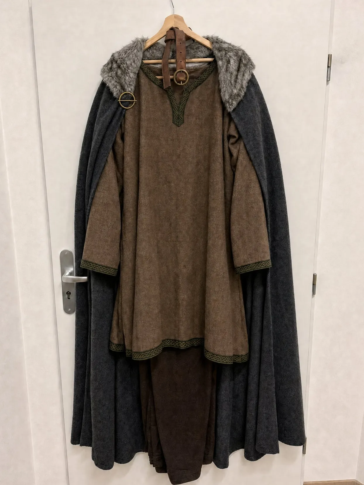 A brown wool tunic with a green woven band, a leather belt and a charcoal cloak with a grey fur collar and a ring brooch, on a hanger on a white door