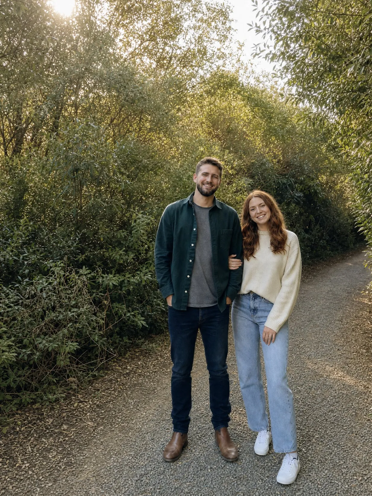 A couple standing on a gravel path between green bushes, he in a dark green overshirt and jeans, she in a cream sweater and light jeans