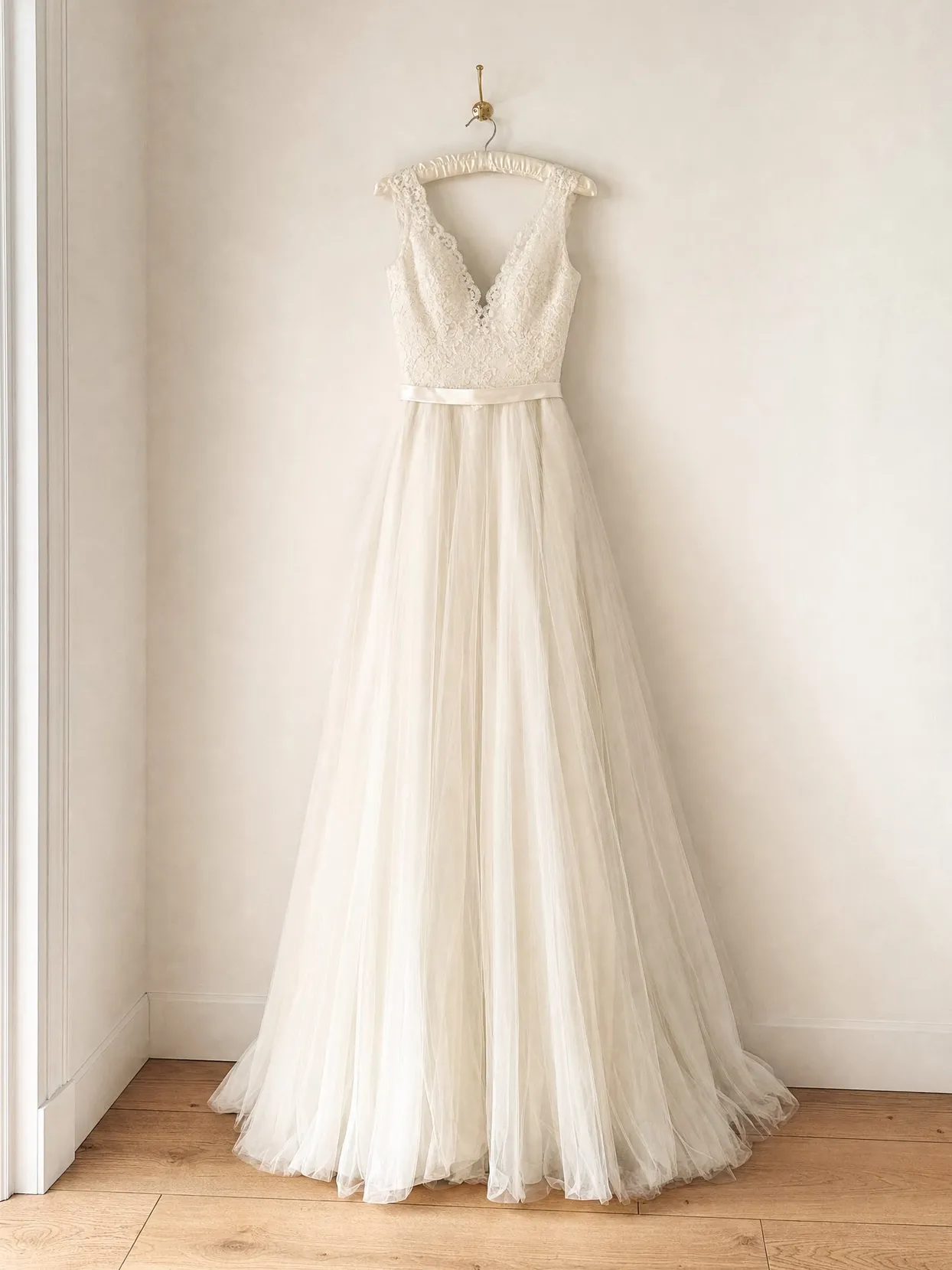 Ein elfenbeinfarbenes Brautkleid aus Spitze und Tüll auf einem Bügel vor einer weißen Wand.