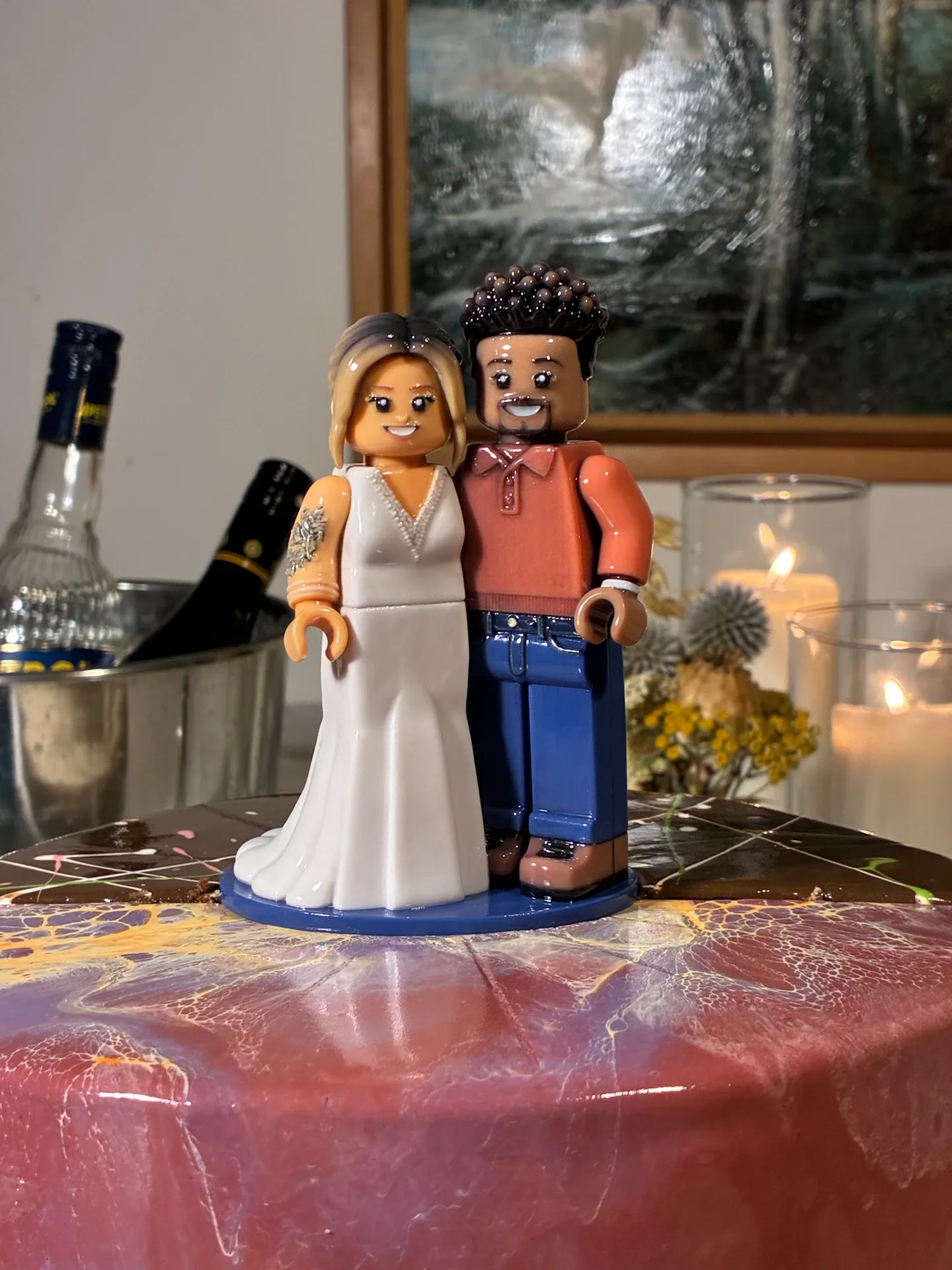 Glossy block-style figure of a bride in a white gown and a groom in a coral shirt and jeans, on a blue base on top of a marbled cake