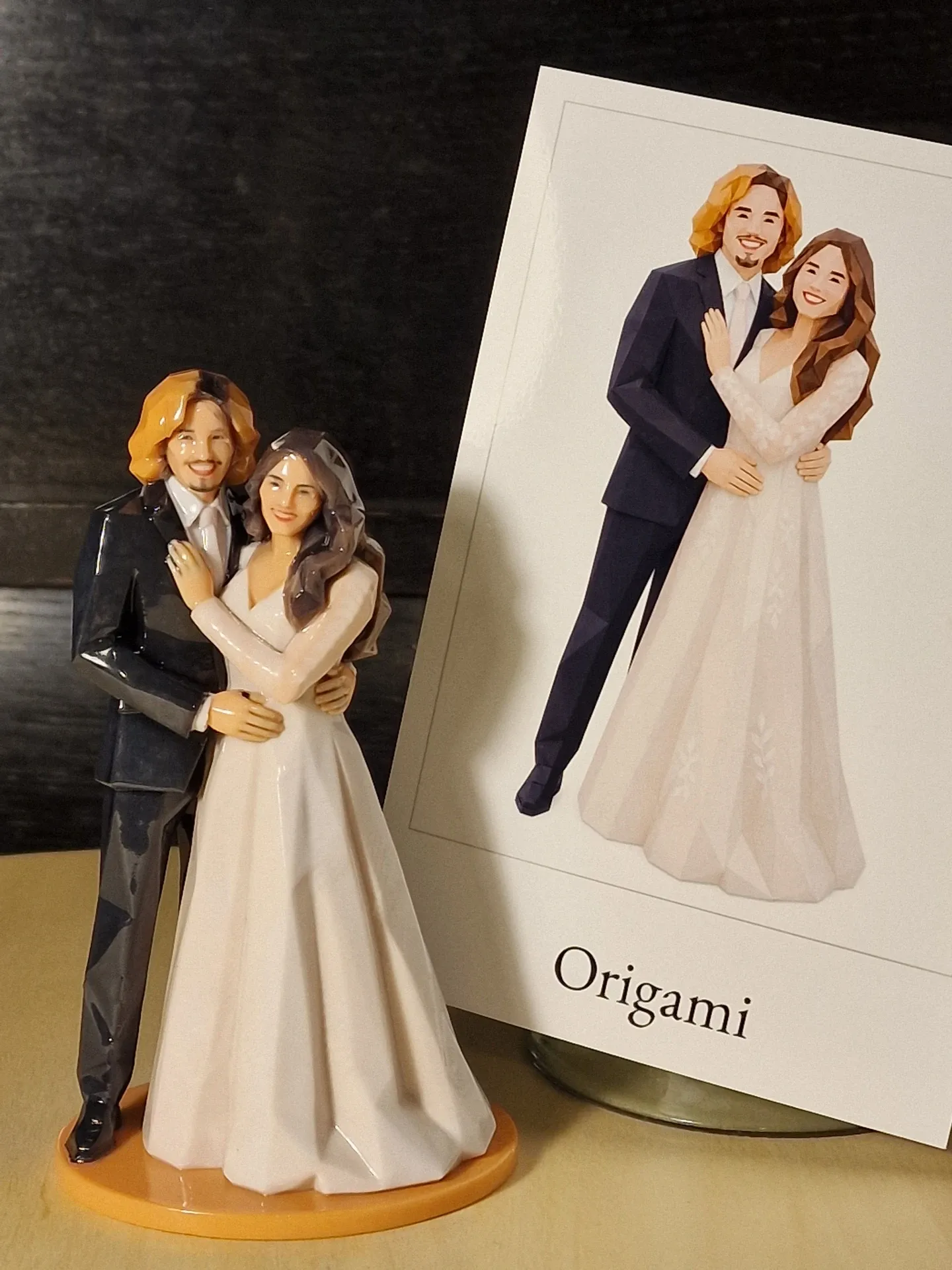 Glossy faceted couple figure, groom in a dark suit and bride in a white gown, on an orange base, beside a printed card of the same design
