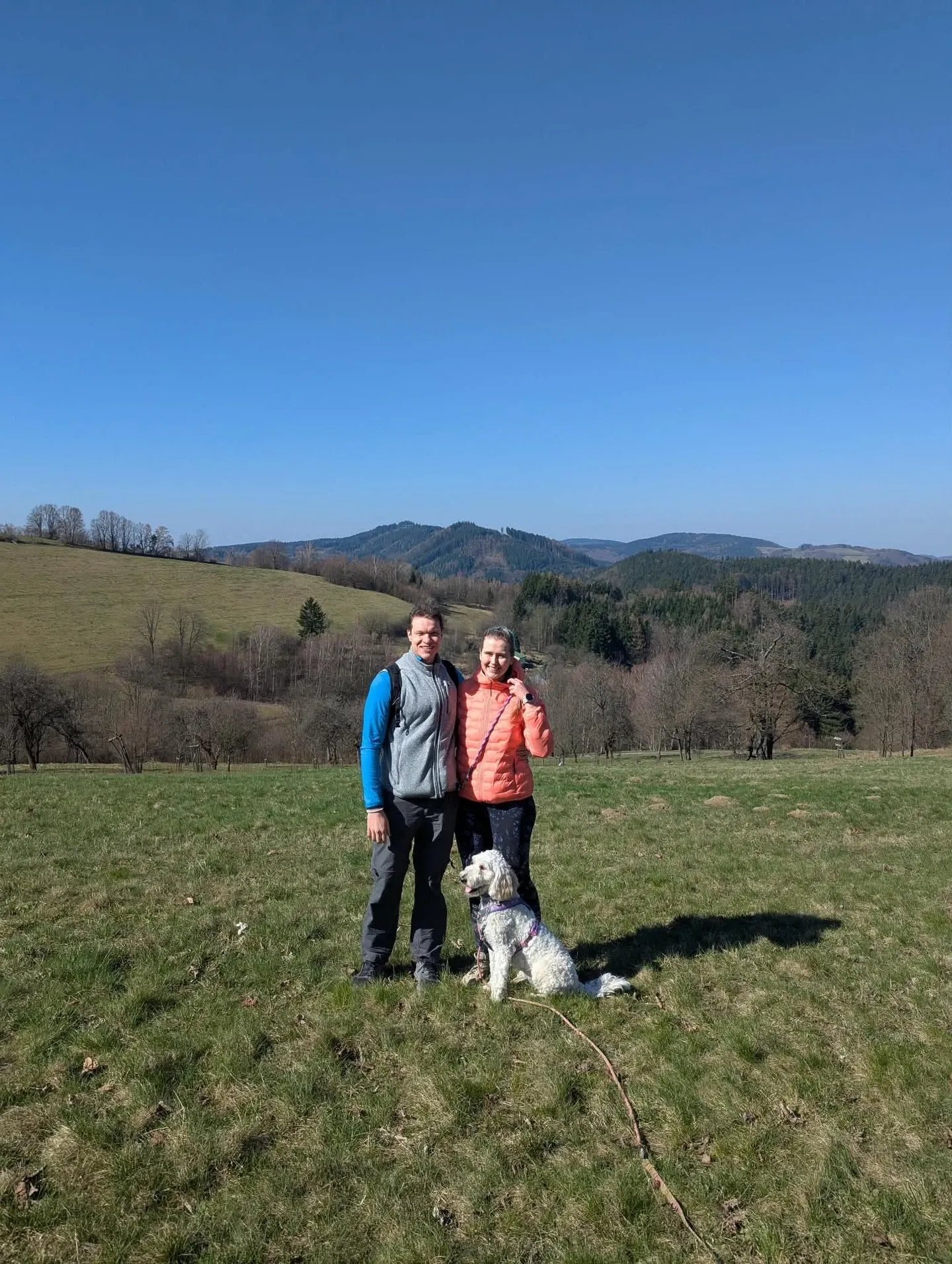 A man and a woman in outdoor jackets stand in a hilly meadow, a white curly dog sitting at their feet