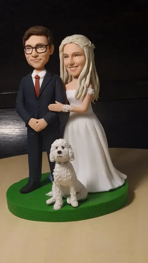 Clay figure of a groom in a navy suit and red tie and a bride in a white gown, with a white curly dog sitting in front, on a green round base