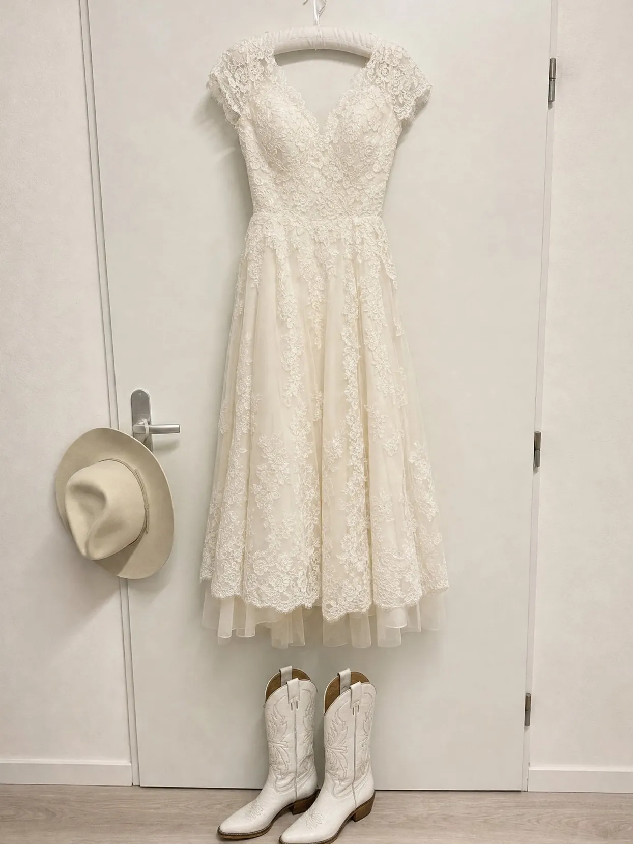 An ivory lace wedding dress hanging on a door, with white cowgirl boots below and a cream hat on the handle.