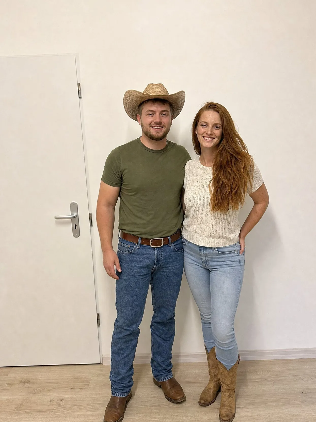 A couple standing against a white wall, he in a straw cowboy hat and jeans, she in jeans and cowboy boots.
