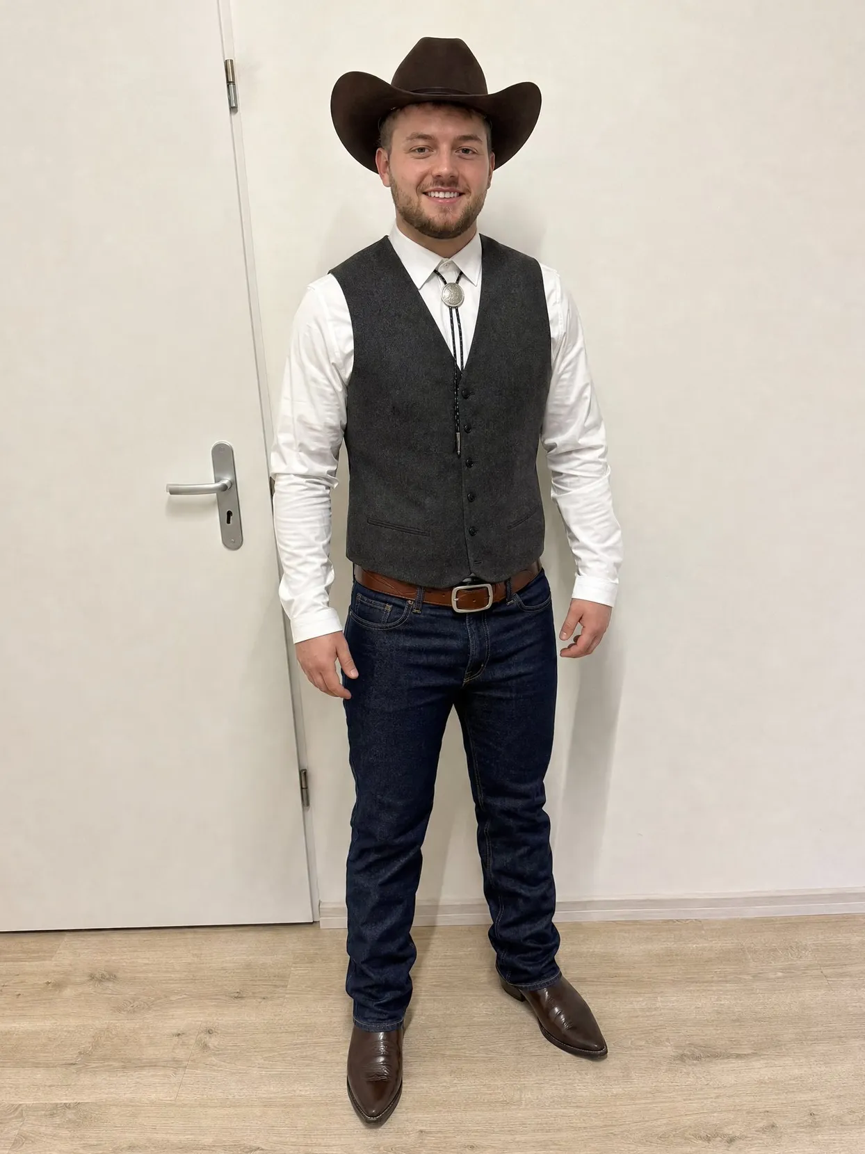 A man in a felt cowboy hat, vest, bolo tie, jeans and cowboy boots standing by a white door.