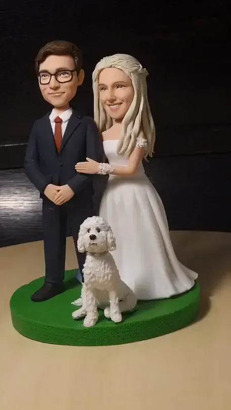 A clay bride and groom topper with a white curly dog at their feet, on a round green base