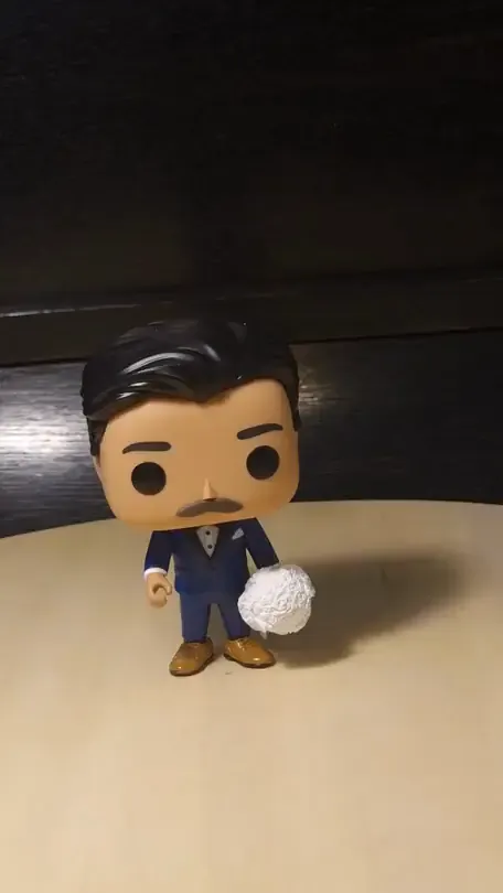 A vinyl toy figure with a large head and a navy suit, holding a white bouquet