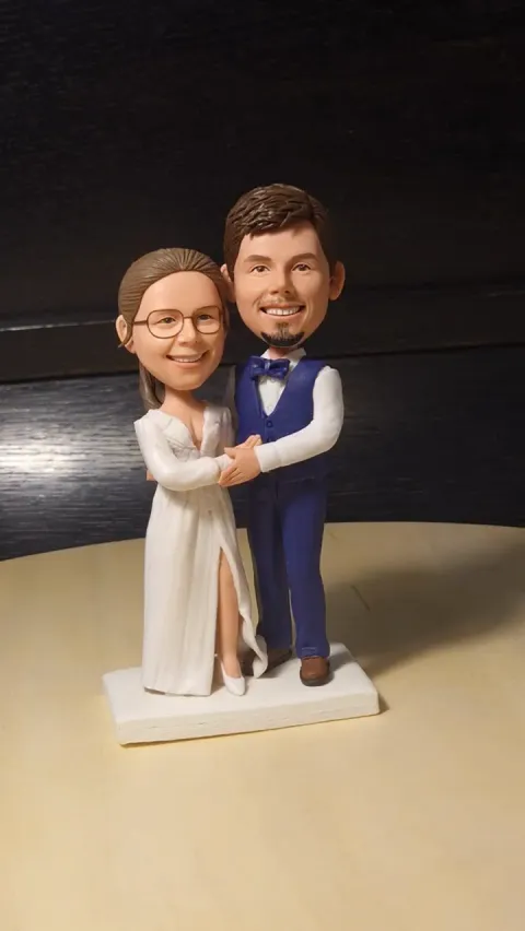 Clay figure of a bride in glasses and a white dress holding hands with a groom in a blue vest and bow tie, on a white base