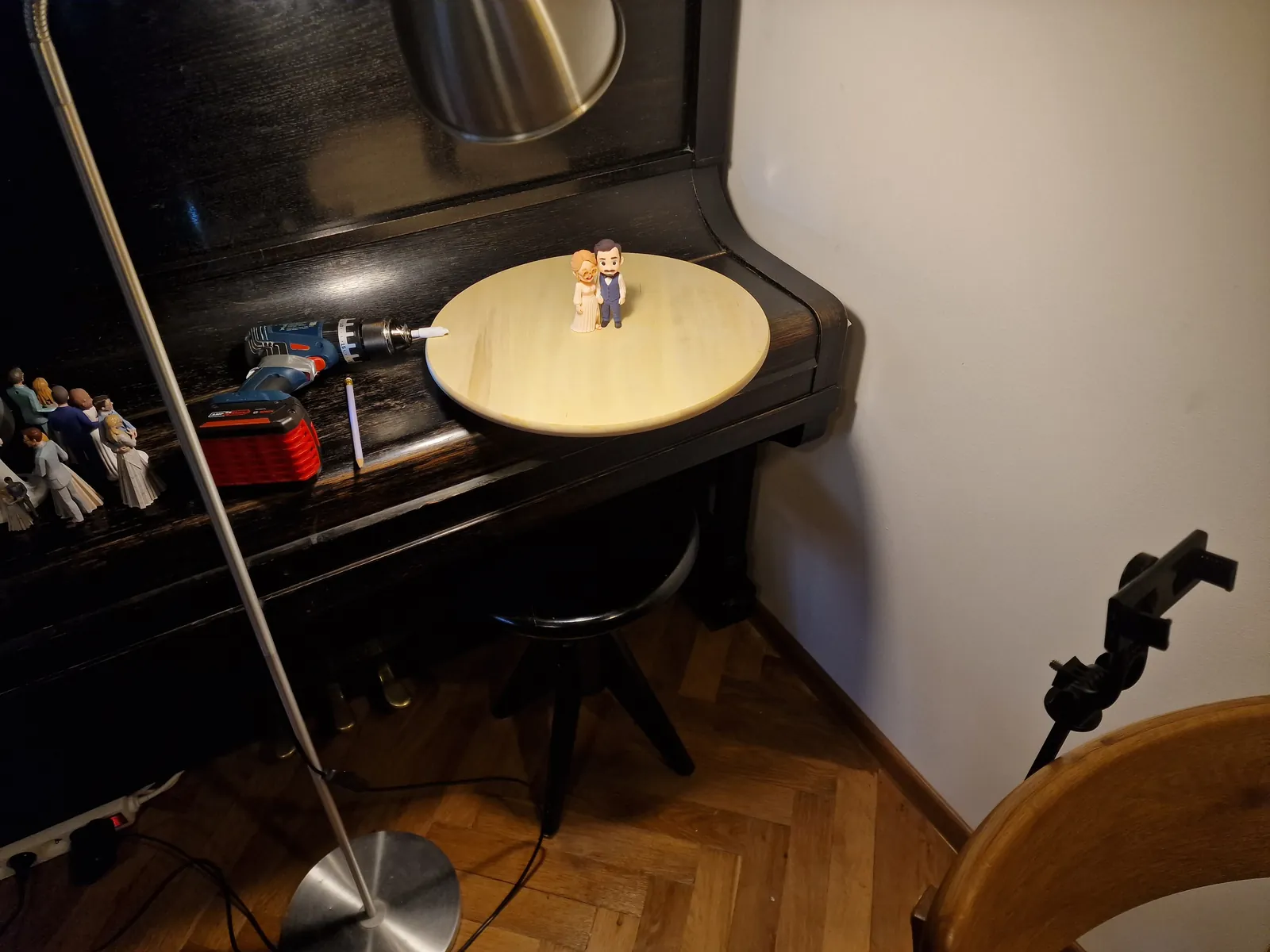 Isang kahoy na disc na hawak ng cordless drill sa ibabaw ng takip ng piano, may nakatayong topper doon, isang lampara sa stand at isang telepono na naka-clamp sa harap